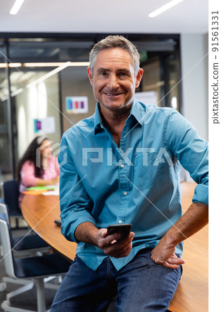 Portrait of smiling mature caucasian businessman holding smart phone while colleagues in background Portrait of smiling mature caucasian businessman holding smart phone while colleagues in background 91561331