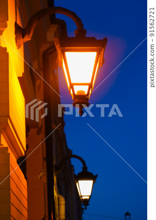 Retro lantern with yellow warm light on the facade of the building. Street lights, illumination. Twilight on the street of the old city. Old street lamp glows in the evening Retro lantern with yellow warm light on the facade of the building. Street lights, illumination. Twilight on the street of the old city. Old street lamp glows in the evening 91562721