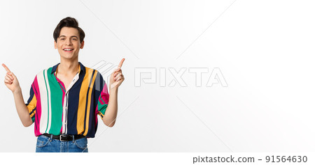 Happy young gay man pointing fingers sideways, showing two choices and smiling, standing against white background Happy young gay man pointing fingers sideways, showing two choices and smiling, standing against white background 91564630