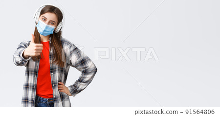 Social distancing, leisure and lifestyle on covid-19 outbreak, coronavirus concept. Pleased cheerful cute girl in medical mask and headphones, showing thumb-up, recommend or approve 91564806