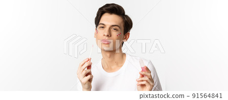 Close-up of beautiful queer person with glitter on face applying lips gloss and looking self-assured at camera, standing over white background 91564841