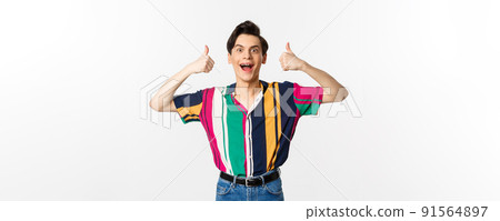 Excited and amazed young man showing thumbs up with impressed expression, standing over white background 91564897