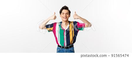 Young cheerful man smiling, showing thumbs up in approval, praise something good, standing over white background Young cheerful man smiling, showing thumbs up in approval, praise something good, standing over white background 91565474
