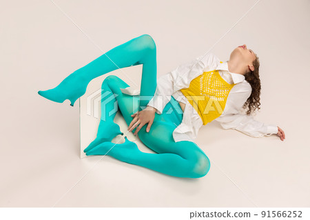 Studio shot of young sad girl in bright cyanic tights posing with mirror isolated over white background. Vivid style, beauty, queer, freak, fashion concept. 91566252