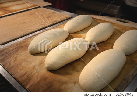 Several raw French rolls lie on a wooden baking sheet in the bakery's bread room before baking in the oven. 91568045