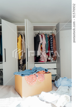 A cardboard box of selected clothes for donating to a Charity shop standing on the bed. Decluttering, Sorting clothes, and Cleaning Up. Reuse, second-hand. Conscious consumer, sustainability. 91570932