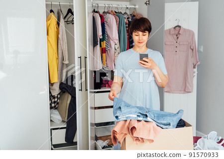 Woman taking photo of the shorts using her smartphone for selling or donating her clothes. Decluttering , Sorting Clothes, And Cleaning Up wardrobe. Reuse, second-hand concept. Conscious consumer. 91570933