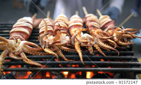 Asian street food in Bangkok - Thailand. Unrecognizable man grilling a squid or seafood barbecue on the charcoal fire wood. 91572147