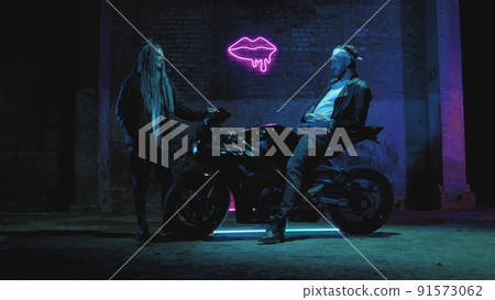 A guy on a super sport motorcycle stands talking to a girl against a pink neon sign A guy on a super sport motorcycle stands talking to a girl against a pink neon sign 91573062