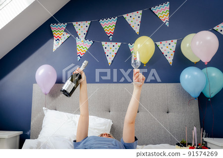 Woman with arms raised holding bottle and glass of champagne lying on the bed with festive cake in the decorated bedroom.Time for yourself Celebration at home. Happy birthday concept. Selective focus. Woman with arms raised holding bottle and glass of champagne lying on the bed with festive cake in the decorated bedroom.Time for yourself Celebration at home. Happy birthday concept. Selective focus. 91574269