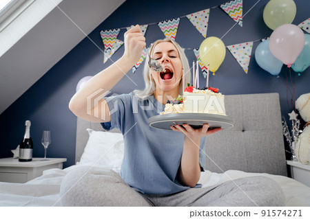 Sad, upset woman in pajama and party cap eating by spoon her birthday cake sitting on the bed in decorated bedroom. Celebrates birthday alone. Jamming of negative emotions. Selective focus. 91574271