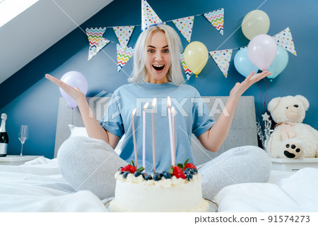 Excited emotional woman in birthday party cap with festive cake with candles on the bed in decorated bedroom. Morning surprise. Happy birthday concept. Selective focus Excited emotional woman in birthday party cap with festive cake with candles on the bed in decorated bedroom. Morning surprise. Happy birthday concept. Selective focus 91574273