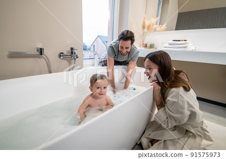 Child looking at camera in bathtub and parents watching 91575973