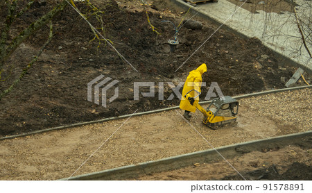 a worker in a yellow raincoat rams the sand before laying asphalt 91578891