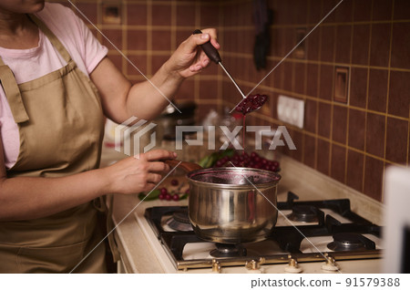 Close-up of a woman, a housewife in an apron, standing at the kitchen stove and making cherry berry jam in metal saucepan in the rusric home kitchen Close-up of a woman, a housewife in an apron, standing at the kitchen stove and making cherry berry jam in metal saucepan in the rusric home kitchen 91579388