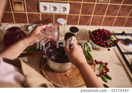 Cropped view of a housewife, woman confectioner pouring delicious freshly brewed cherry berry confiture into sterilized glass jar at home kitchen. Cropped view of a housewife, woman confectioner pouring delicious freshly brewed cherry berry confiture into sterilized glass jar at home kitchen. 91579397