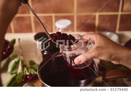 Confectioner's hands pouring freshly made cherry berry confiture into sterilized jars at rustic home kitchen. Preparing homemade preserves for the winter 91579400