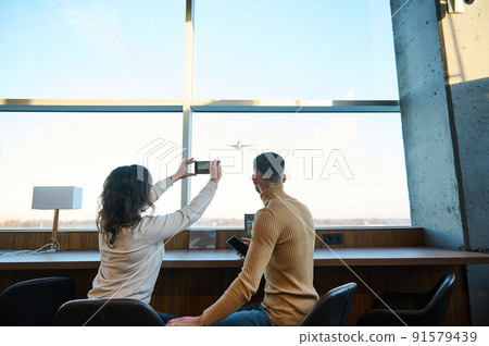 Rear view of a couple taking pictures of an airplane taking off against sunset sky in the airport departure terminal, waiting to board the flight 91579439