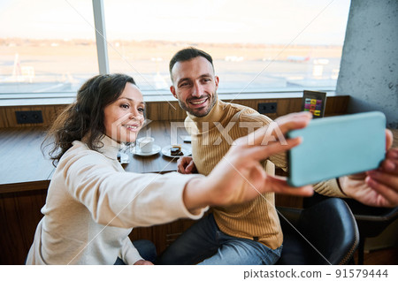 Beautiful couple in love holds smartphone in outstretched hands and taking selfie while enjoying breakfast together in the airport departure terminal Beautiful couple in love holds smartphone in outstretched hands and taking selfie while enjoying breakfast together in the airport departure terminal 91579444