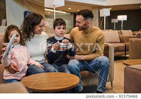 Portrait of happy family in VIP lounge of the airport departure terminal waiting to board the flight. Children - boy and girl playing with toy planes Portrait of happy family in VIP lounge of the airport departure terminal waiting to board the flight. Children - boy and girl playing with toy planes 91579466