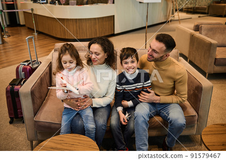 Loving mom and dad tenderly hug their kids playing with toy airplanes as they wait to board their flight in VIP lounge of airport departure terminal. Loving mom and dad tenderly hug their kids playing with toy airplanes as they wait to board their flight in VIP lounge of airport departure terminal. 91579467