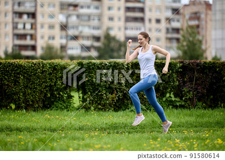 Young woman in tank top and leggings jogging in public park next to residential area with high-rise apartment buildings. Young woman in tank top and leggings jogging in public park next to residential area with high-rise apartment buildings. 91580614