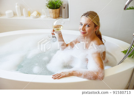 Attractive young blonde female sitting in bathtub with foam and holding white wine glass. Attractive young blonde female sitting in bathtub with foam and holding white wine glass. 91580713