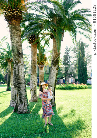 Mom holds a little girl in her arms, standing under palm trees on a green lawn 91581128