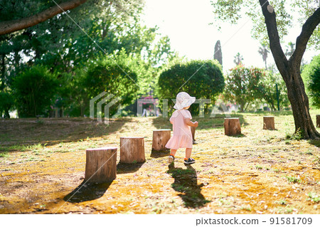 Little girl in a hat walks along the lawn past the stumps. Back view Little girl in a hat walks along the lawn past the stumps. Back view 91581709