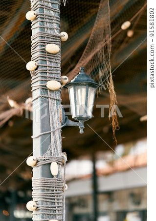 Lantern hangs on a pole braided with fishing tackle under a canopy from which a fishing net hangs Lantern hangs on a pole braided with fishing tackle under a canopy from which a fishing net hangs 91581802