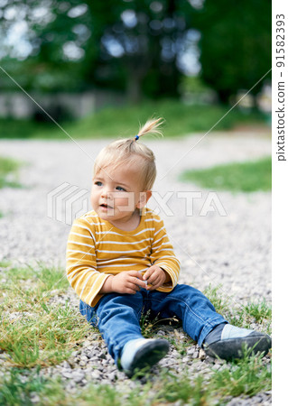 Little girl sits on a gravel path and holds a small pebble in her hand 91582393