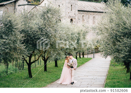 Newlyweds stand on a path in a blooming olive grove against the background of an old building on Lake Como 91582606