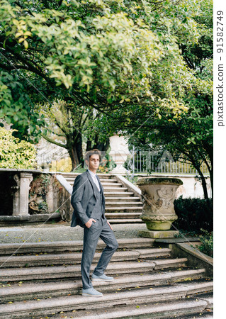 Serious groom stands with his hand in his pocket on the stairs in the park. Lake Como 91582749