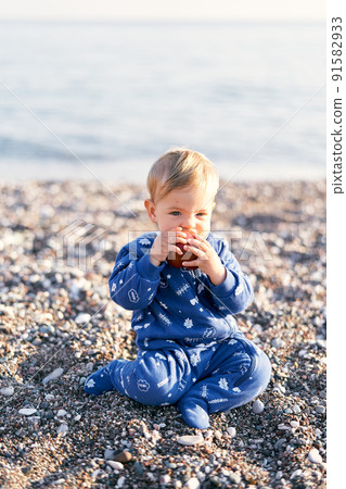 Small child in a blue overalls sits on a pebble beach and eats an apple. Close-up Small child in a blue overalls sits on a pebble beach and eats an apple. Close-up 91582933