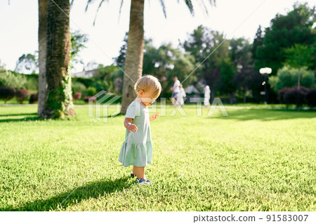 Little girl stands on a green lawn and looks at her feet. Side view Little girl stands on a green lawn and looks at her feet. Side view 91583007