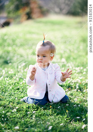 Little toddler with a chamomile behind her ear sits on a green lawn among white flowers 91583014