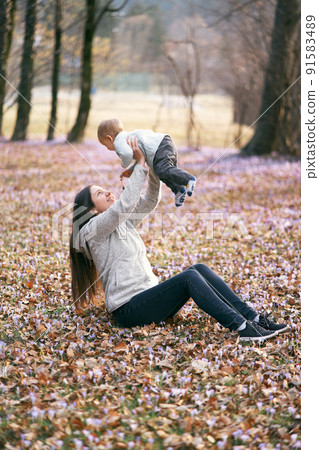 Mom sits on fallen leaves in the park, lifting the baby above her in her arms Mom sits on fallen leaves in the park, lifting the baby above her in her arms 91583489
