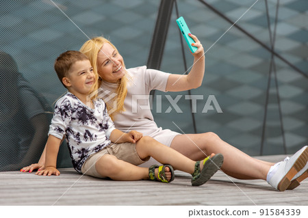 Happy blond woman and little boy sitting on terrace and making selfie on smartphone. Mother and son enjoy time together. Positive young mom spending time with her cute child. Having fun. Family. Happy blond woman and little boy sitting on terrace and making selfie on smartphone. Mother and son enjoy time together. Positive young mom spending time with her cute child. Having fun. Family. 91584339