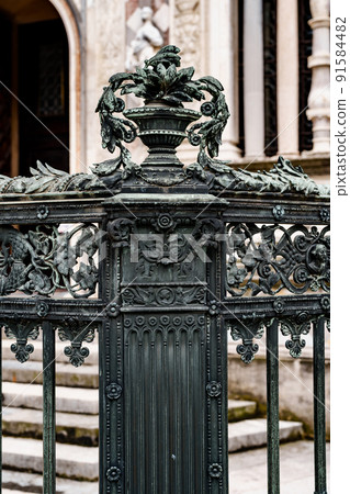 Metal vase with flowers on the pedestal of an old wrought-iron fence Metal vase with flowers on the pedestal of an old wrought-iron fence 91584482