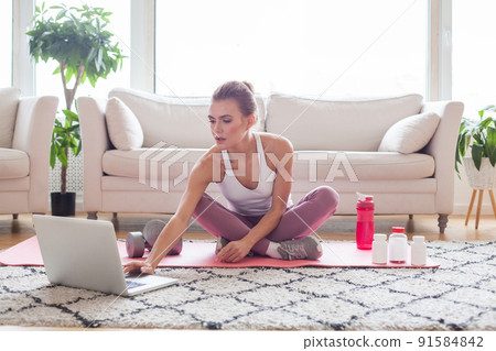 Sporty woman in white and pink sportswear sitting on the floor with dumbbells and a protein shake 91584842