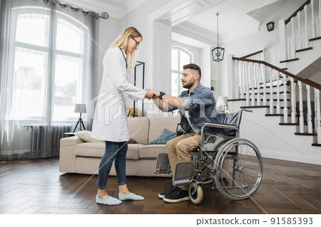 Therapist assisting man to stand up from wheelchair at home 91585393