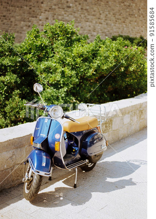 Blue motor scooter stands on paving stones near a stone fence Blue motor scooter stands on paving stones near a stone fence 91585684