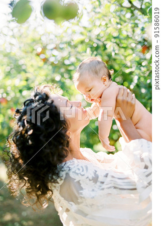 Woman in a white dress raises a baby in her arms in the garden 91586019
