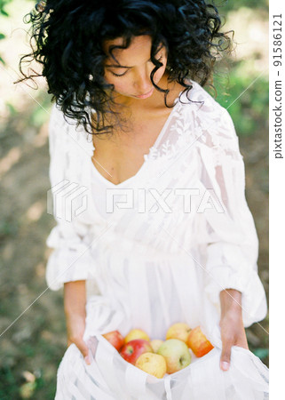 Woman in a white dress holds pile of apples in her hem Woman in a white dress holds pile of apples in her hem 91586121