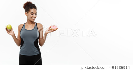 Healthy and diet concept - Beautiful sporty African American make a decision between donut and green apple. Isolated on white background. Healthy and diet concept - Beautiful sporty African American make a decision between donut and green apple. Isolated on white background. 91586914