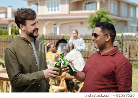 Happy young interracial buddies clinking with bottles of beer at picnic Happy young interracial buddies clinking with bottles of beer at picnic 91587526