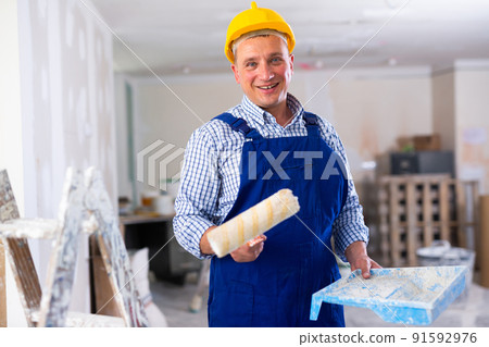 Portrait of positive man painter in a protective helmet and overal with paint roller Portrait of positive man painter in a protective helmet and overal with paint roller 91592976