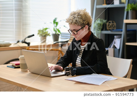 Young woman with prosthetic arm typing on laptop working at her workplace at office 91594773