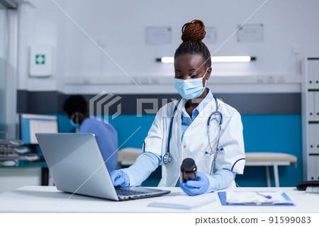 Healthcare specialist medic holding vial of medicament while using laptop to analyze treatment results. Doctor examining medicine side effects and benefits while wearing virus protection facemask. Healthcare specialist medic holding vial of medicament while using laptop to analyze treatment results. Doctor examining medicine side effects and benefits while wearing virus protection facemask. 91599083