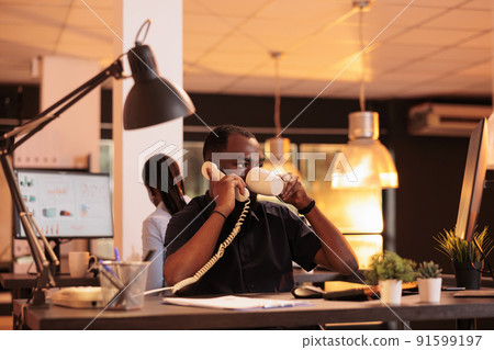 Startup employee using landline phone to have remote conversation during sunset. Chatting on office telephone with colleagues to work on business report and do paperwork contract. 91599197